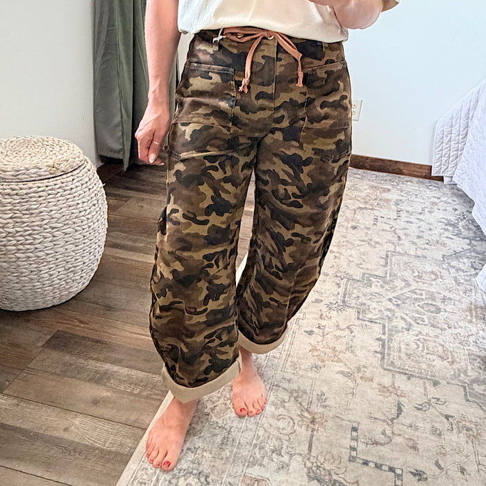A person wearing loose-fit camouflage pants with a tan drawstring waist, standing barefoot on a patterned rug in a bedroom setting.