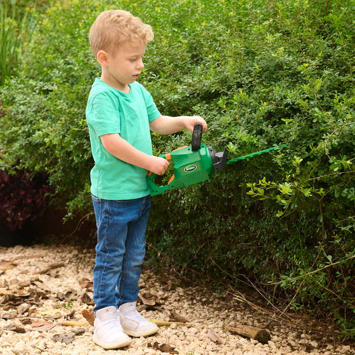 A young boy in a green shirt and jeans trims a bush with the Red Toolbox Scotts Battery-Operated Hedge Trimmer, standing on a garden path—perfect for safe, realistic yard play.