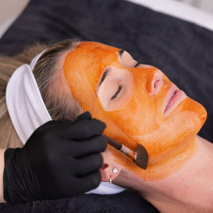 Person receiving an orange facial mask treatment with a gloved hand applying it.