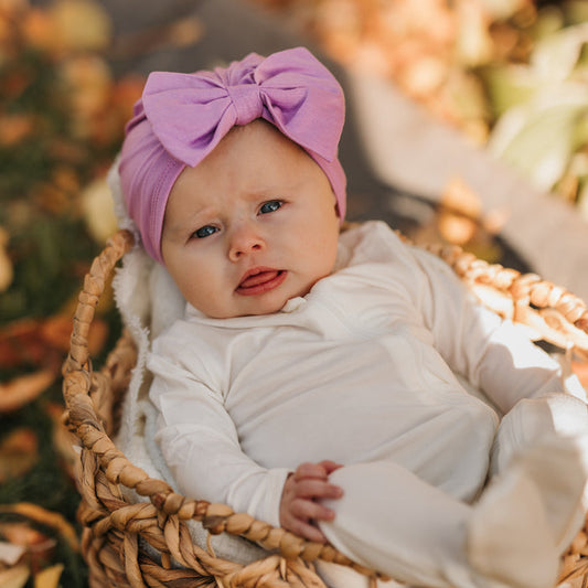 Infant laying in a wicker basket wearing the Bow Headwrap in Poi and white footie