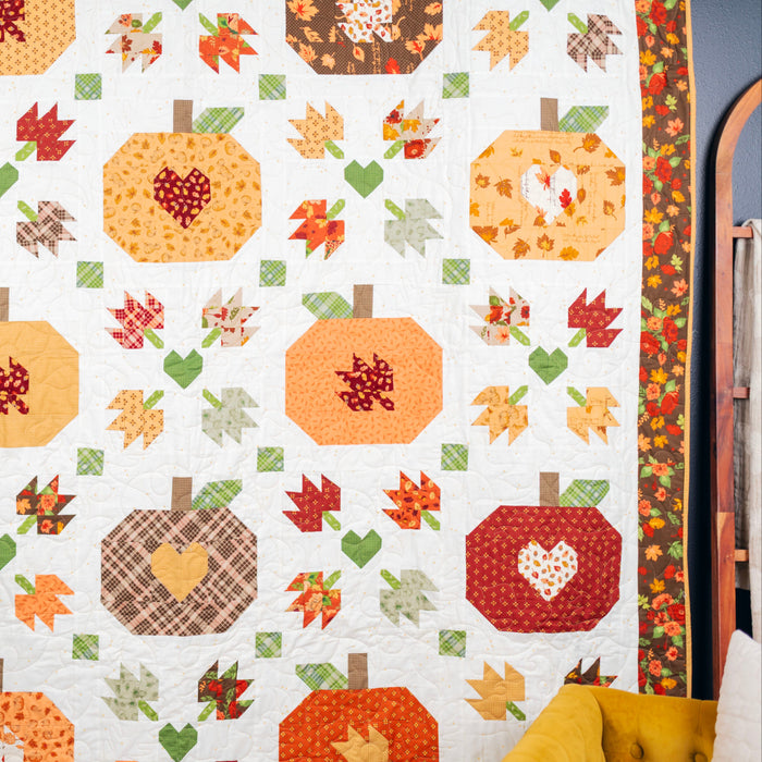 A handmade autumn-themed quilt hanging on a wall, featuring stitched pumpkins, leaves, and hearts in warm fall colors like orange, red, yellow, green, and brown on an off-white background.