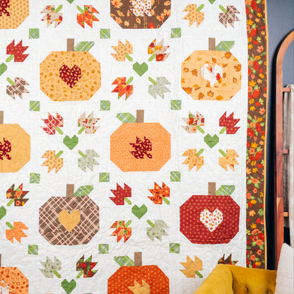 A handmade autumn-themed quilt hanging on a wall, featuring stitched pumpkins, leaves, and hearts in warm fall colors like orange, red, yellow, green, and brown on an off-white background.