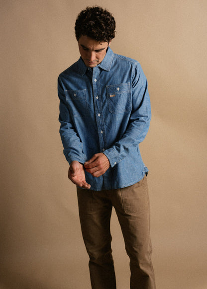 Man in a blue denim shirt adjusts sleeve, wearing brown pants, against a beige background, looking down thoughtfully.