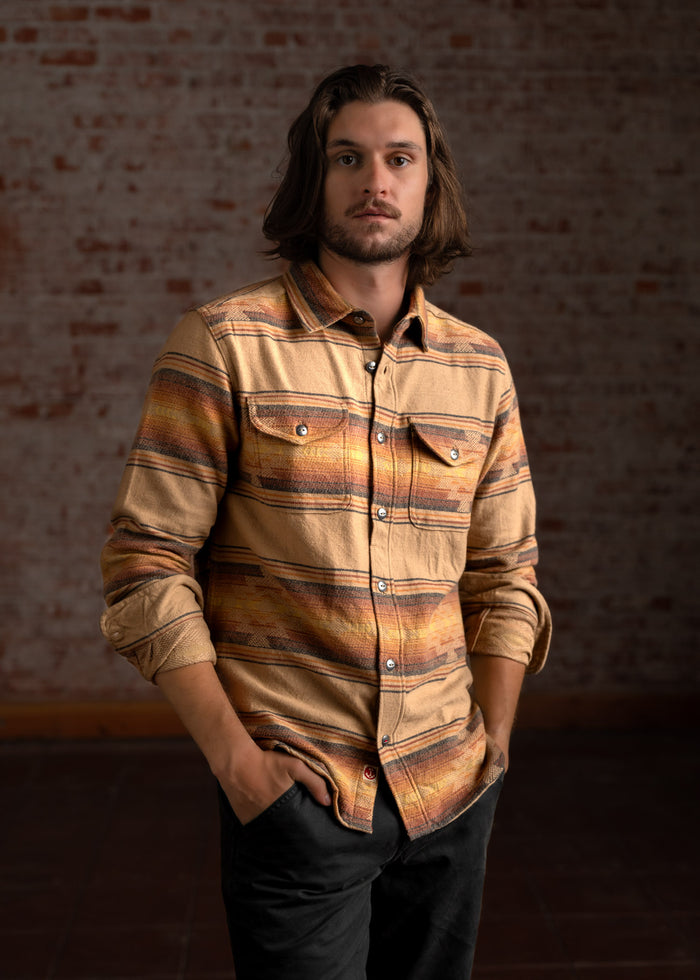 Man with long hair in a patterned shirt stands confidently against a brick wall, hands in pockets, exuding a relaxed vibe.