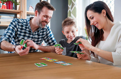 A smiling family of three plays Mattel Games Skip-Bo Junior by Mattel at a table, happily engaged with cards in a bright, cozy room, enjoying this fun, kid-friendly card game together.
