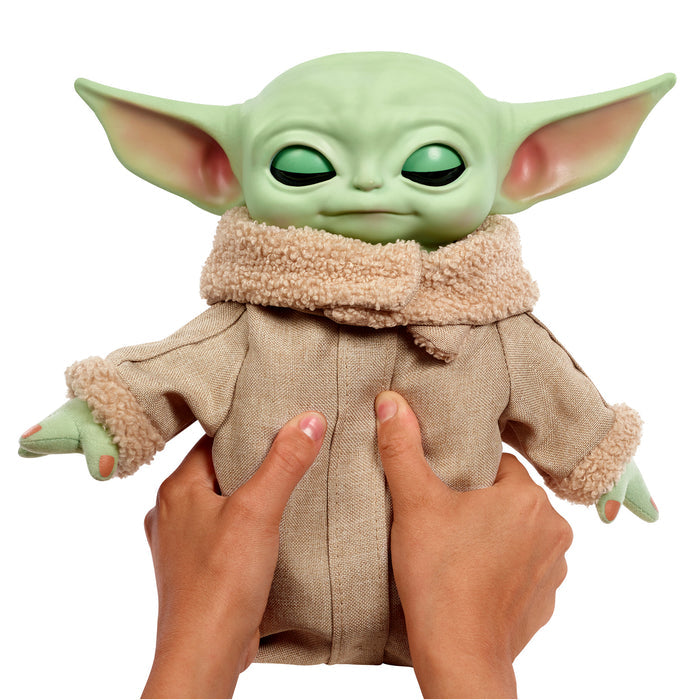 A pair of hands holds a Mattel Star Wars Grogu Squeeze & Blink plush, featuring a tan coat with fuzzy collar and cuffs, large green ears, and closed eyes—an adorable gift for any Star Wars fan.