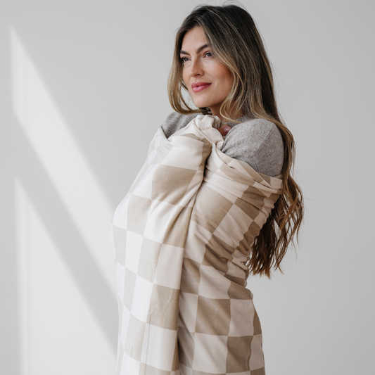 A woman is wrapped in a tan and cream checkered minky stretch blanket. This Saranoni blanket is very stretchy and durable. This soft blanket is perfect for relaxing, gifting, and taking on the go. It is also well made to last you for years to come.