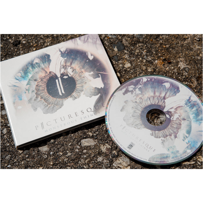 Faded white CD case with a human eye faded onto the background.  There is text at the bottom that reads Picturesque and Monstrous Things. Next to the cover is a cd all of which is laying on top of a road.