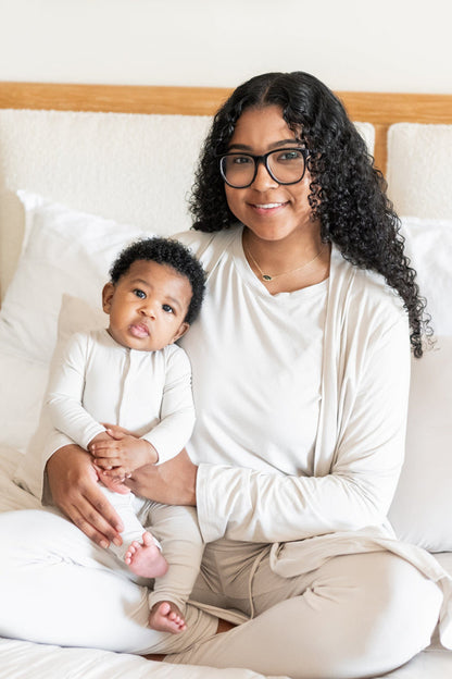 Mother and baby sitting on a bed matching while baby wears a Kyte Baby Snap Romper in Oat