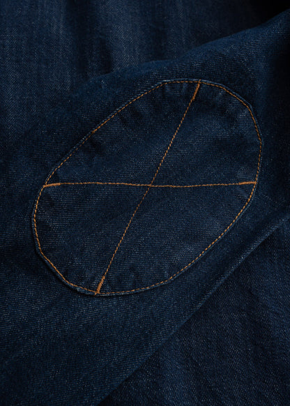 Close-up of a denim jacket's elbow patch with orange stitching, forming an X pattern on dark blue fabric.