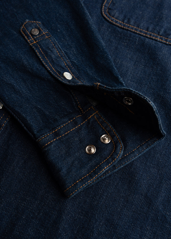 Close-up of a dark denim shirt with visible orange stitching and metal snap buttons, showcasing detailed craftsmanship.