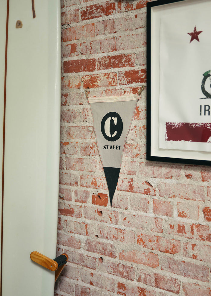 A surfboard leans against a brick wall with a "C Street" pennant and framed art, creating a coastal, vintage vibe.