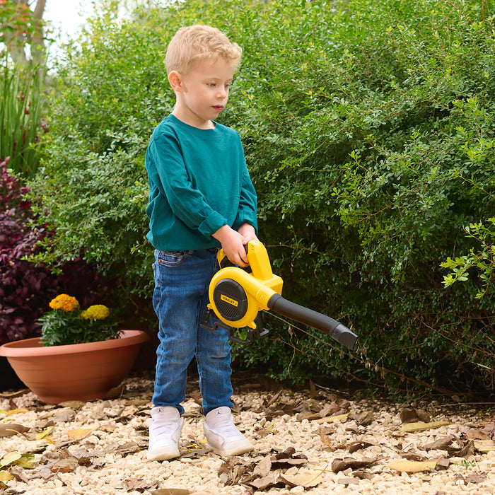 A young boy with blond hair, in a green shirt and jeans, uses the Red Toolbox Stanley Jr. Power Blower on a gravel garden path, with green bushes and yellow flowers in the background of this playful outdoor scene.