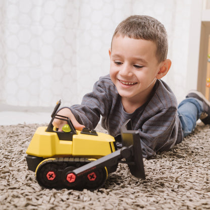 A young boy smiles on the carpet while playing with his Red Toolbox Stanley Jr. Take Apart XL Bulldozer, enjoying hands-on construction fun.
