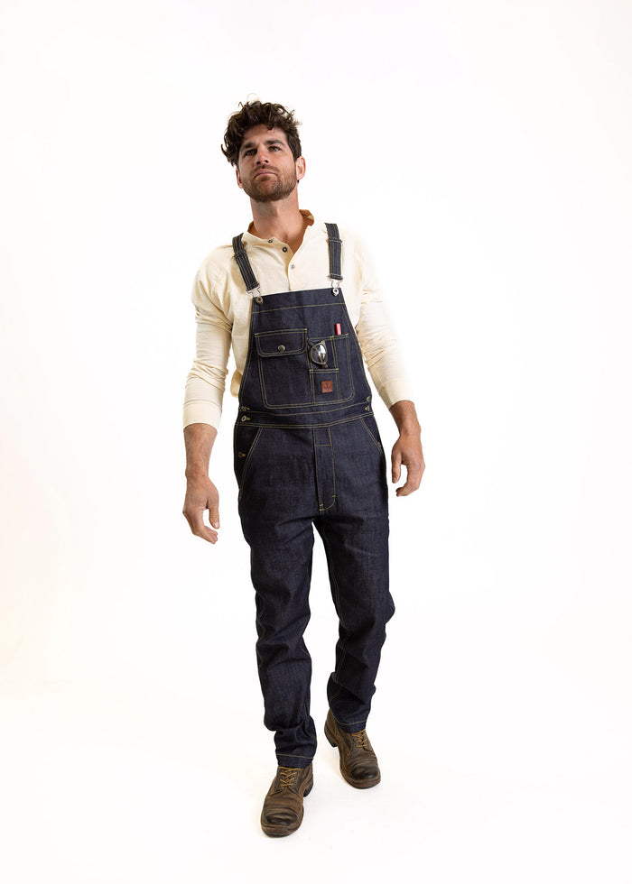 Man in denim overalls and boots stands confidently against a white background, wearing a cream shirt.