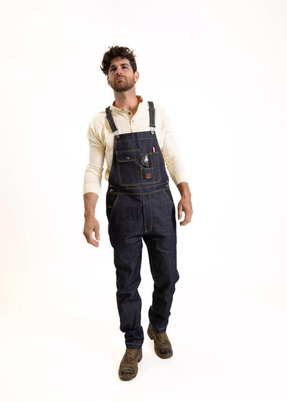 Man in denim overalls and boots stands confidently against a white background, wearing a cream shirt.