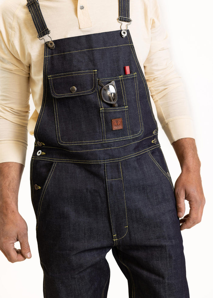 Man in denim overalls with yellow stitching, glasses in pocket, wearing a beige shirt, hands relaxed by sides.