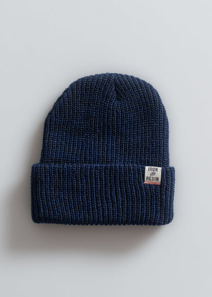 A navy blue knit beanie with a folded brim, featuring an "Iron and Resin" label, lies on a light gray background.
